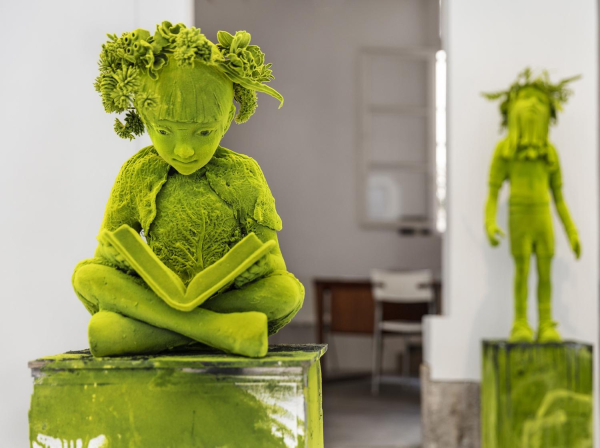 Sculpture of a childlike figure sitting cross-legged reading a book, entirely in a bright green hue, with leafy forms sprouting from their head and chest