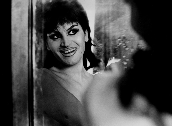 Black and white photograph of a smiling woman with dark shaggy hair and bold makeup looking in a water-sprayed mirror