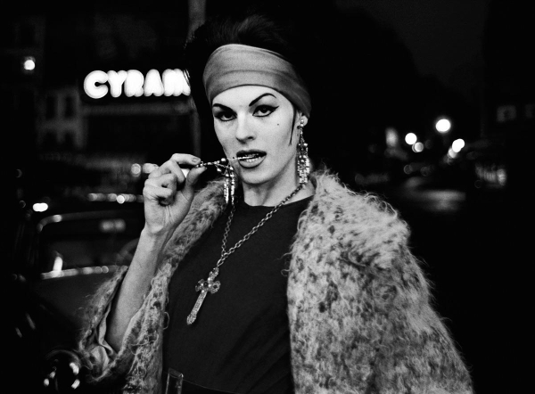 Black and white photograph of a woman with dark hair and bold makeup standing outside at night wearing a big fur coat and heavy cross necklace, holding a piece of jewelry in her mouth and staring suggestively at the camera