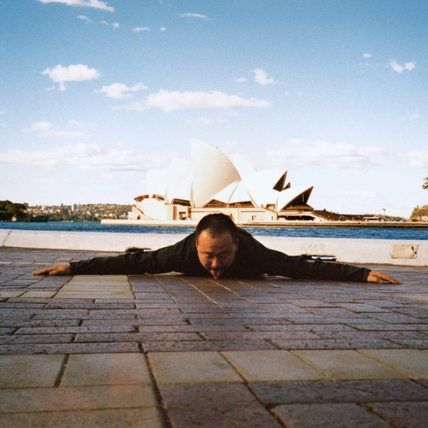 Photo of the artist in Sydney lying prostrate on the ground with his tongue licking the pavement, in front of the Sydney Opera House