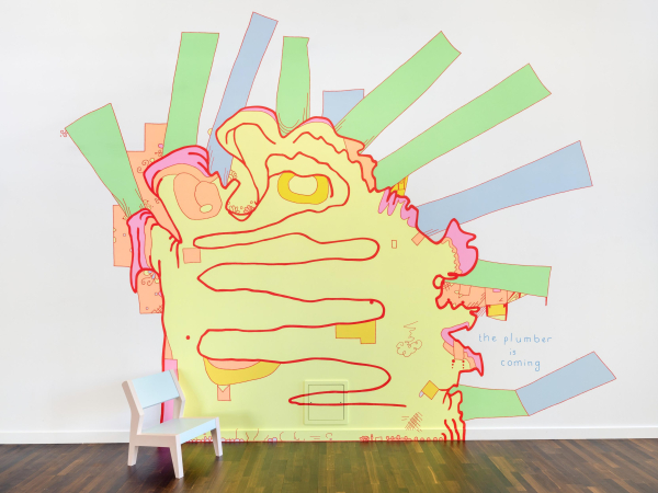 Wall painting of a large bright yellow blobby shape with green and blue rectangles radiating from its edges and the text "the plumber is coming" handwritten next to it. There is a small white chair sitting on the floor in front of the painting.