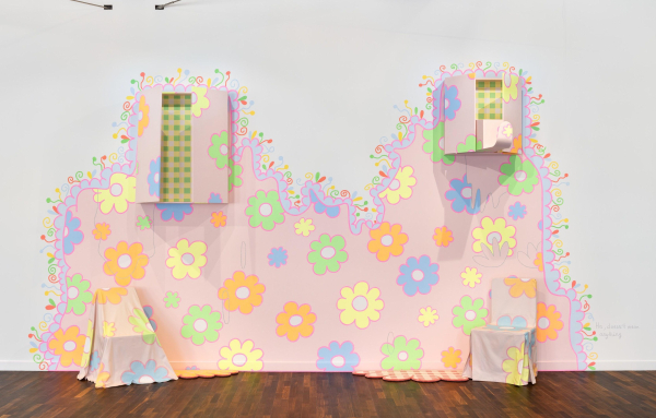 Wall painting of a large pink abstract form decorated with colorful cartoon flowers in bright colors with the words "ha, doesn't mean anything" handwritten next to it. There are painted windows built into the wall within the abstract shape, as well as two chairs on the floor covered in fabric decorated in the same floral pattern