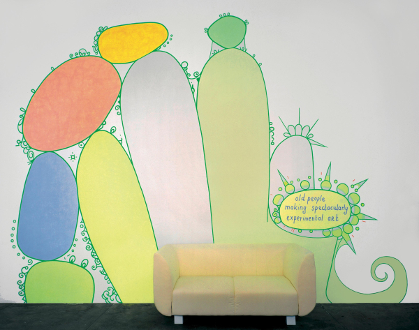 Wall painting of a large cactus-like formation in bright colors with the words "old people making spectacularly experimental art" handwritten in a text bubble. There is a yellow couch sitting on the floor in front of the painting.