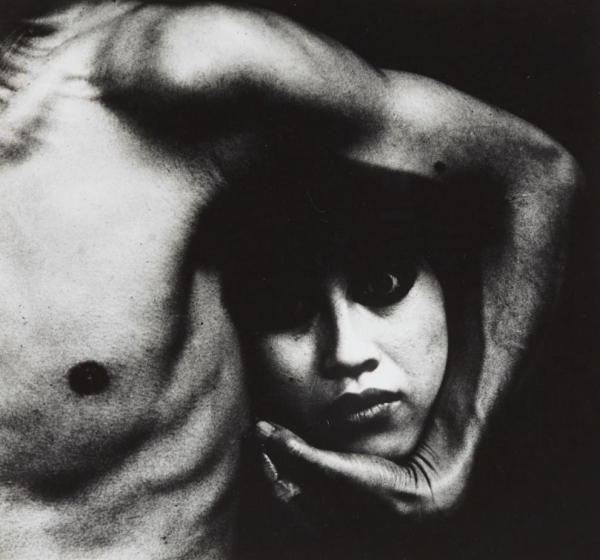 Black and white photo of a shirtless man, with only his torso and left arm visible in frame. A shocked woman's head is cradled under his arm