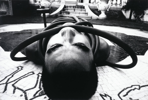 Black and white photo of a man with buzzed hair lying down on tiled floor wrapped in black tubing, positioned with his head in the foreground so the viewer is almost eye-level with him