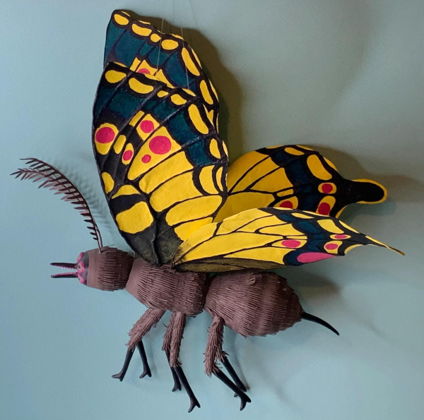 Small sculpture of a butterfly creature seen in profile with a three-part black body and little black boots on its six feet, and bright yellow, blue, and red wings