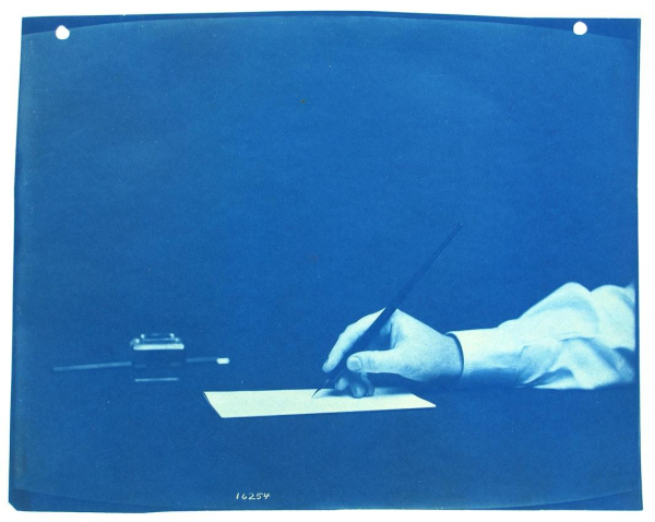 Blue-tinted photo of a hand holding an ink pen over a sheet of paper and a dark desk