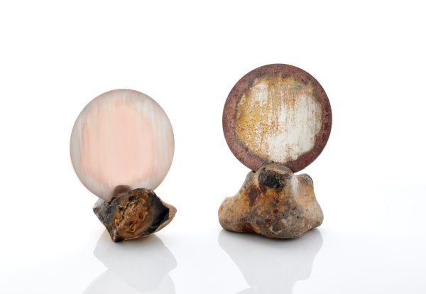 Two round flat metal discs held up vertically in rock stands; one is white and pearlescent, the other is covered in a brown and orange patina