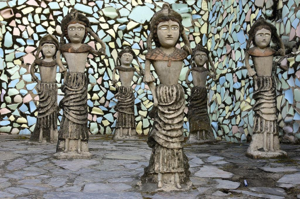 Figures made of grey and brown stone wearing long skirts, each posing with one arm up and the other on their hips as if dancing, against a wall decorated with colorful stone tile