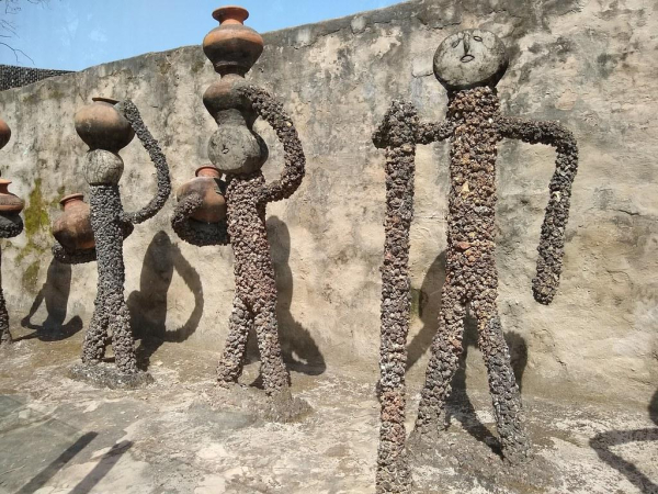 Figures made of grey and brown stone, some holding pottery vessels, against a flat, light brown wall