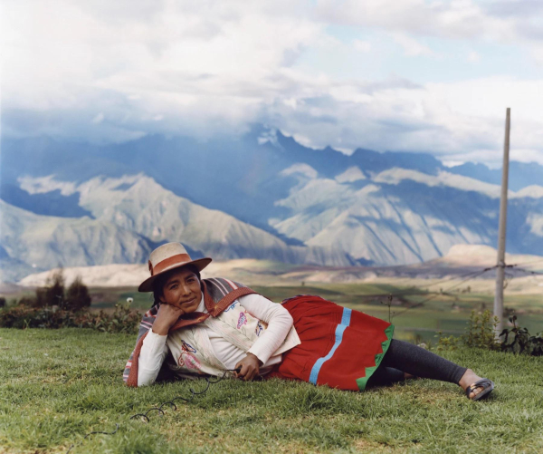 Photo of an indigenous Peruvian woman wearing a red skirt, white top and shawl, and hat, reclining on her side holding her head in her right hand. Behind her is a broad snowy mountain landscape and hovering clouds