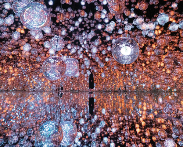 A large space with video projections showing hanging purple orchids and greenery along the floor and walls of a gallery. Two figures in white are in the center of the room looking up