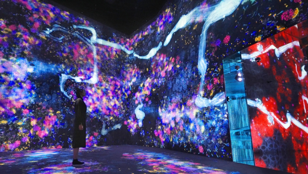 A large space with video projections showing streaks of bright light and purple starburst motifs along the floor and walls of a gallery. A figure wearing a black dress stands in the center of the room