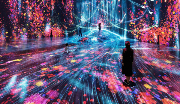 A large space with video projections showing streaks of bright light and pink and orange floral motifs along the floor and walls of a gallery. Some visitors stand around the room.