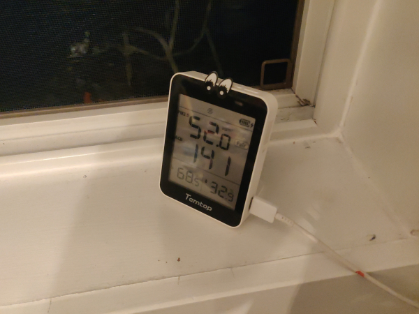 An AQI monitor on a window sill next to an open window. It shows pm2.5 at 52.0, with an AQI of 141.