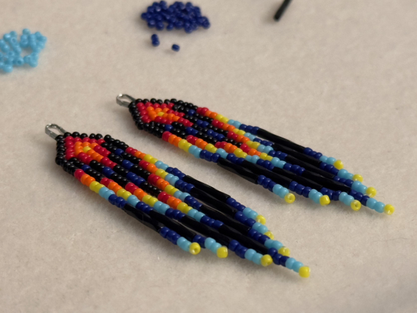 A pair of black, red, orange, yellow, and blue mini beaded fringe earrings on a bead mat