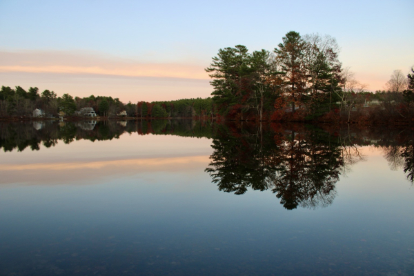 A tranquil lake scene at dusk, featuring trees reflecting in the calm water. The shore includes a few houses, surrounded by a mix of evergreen and autumn-colored foliage. The sky shows soft pastel hues of sunset.