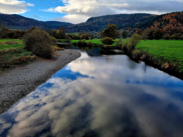 River Conwy still reflecting the clouds, stone bank left grass right mountains in the background.