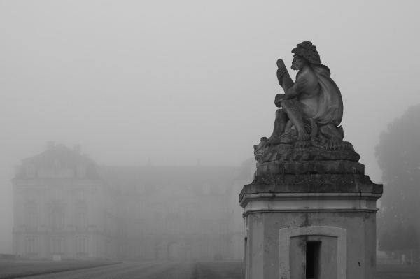 das schwarzweißbild im querformat zeigt die statue einer laute spielenden figur, die auf einen hohen socken sitzt.
im hintergrund im dichten nebel ist die ostfassade des schlosses augustusburg in brühl zu sehen.