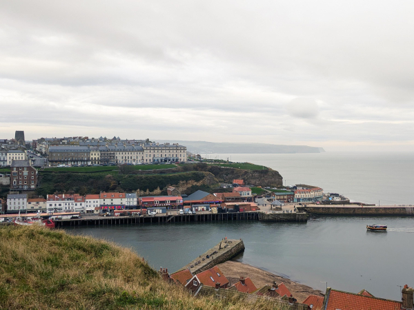 View of Whitby from the graveyard at St. Mary's Church, one of the major landmarks in Bram Stoker's Dracula. Photo is taken from near the grassy cliff edge, looking down over nearby houses to the harbour below. Across the water is the other side of the harbour with pleasingly old classy buildings at the top of the cliffs and some very tacky shops and arcades along the bottom.