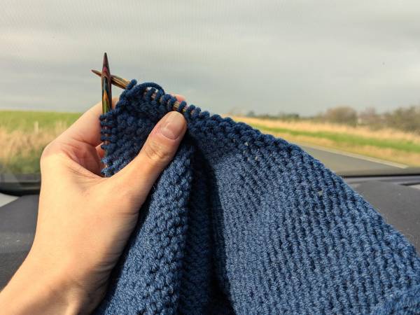 Holding up a navy blue stockinette knitting project in front of the car window. It looks like a grey and featureless day outside.