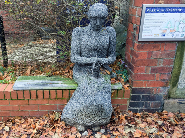 A sculpture of a woman sitting on a bench. She's wearing what looks like a long, old-fashioned high-collared dress, and is knitting a gansey sweater. The whole thing seems to be made out of some kind of dense jumbled wire. Beside her is a sign titled "walk with heritage" with more information about ganseys.