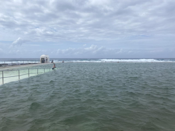 Photo looking across the main ocean pool at Merewether Ocean Baths and out to sea on a cloudy day. The water is such a light green it's almost white over the entry ramp on the left; the rest of the water in the pool is a deeper grey-green. Over the back wall, the sea is more of a dark blue, with an edge of white where the waves are breaking just behind the Baths.