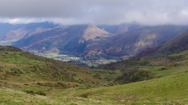 La vue vers le val d'Azun et le village d'Arrens-Marsous depuis le col du soulor. Le plafond nuageux est assez bas et accroche des montagnes de taille intermediaire, mais il y a de beaux eclairages de soleil sur certaines pentes aux couleurs d'automnes, et sur le village et la vallée. Au 1er plan, des collines d prairies avec des fougeres cramoisies. Teintes pastel vert, rouge-pourpre, brun.