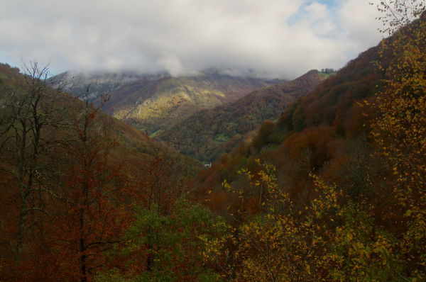 Vue plongeant depuis un lacet de la route du col du soulor vers la vallée et Arbeost, montagnes éclairées par endroits entre les nuages, couleurs chatoyantes d'automne voire brulées, rouges intense.