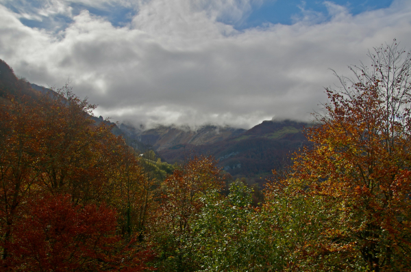 Vue de bord de route vers le col du Soulor et le cirque du Litor. Soleil intense sur les couleurs d'automne des arbres et le toit d'une ferme  surun versant, nuages et quelques trouées de ciel bleu. Lumières intenses de fin de matinée.