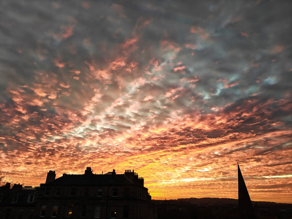 A photograph of the evening sky, with "mackerel" clouds tinged orange, pink and red. At the bottom of the photograph is a silhouette Georgian tenement building and to the right, the silhouette of a church spire.