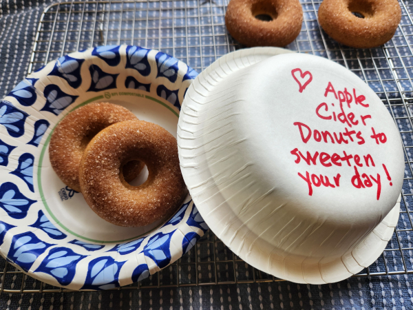 Two apple cider donuts in a paper bowl to deliver to a friend. The bottom of the bowl I use as a lid says apple cider donuts to sweeten your day. The donuts are dipped in cinnamon sugar.