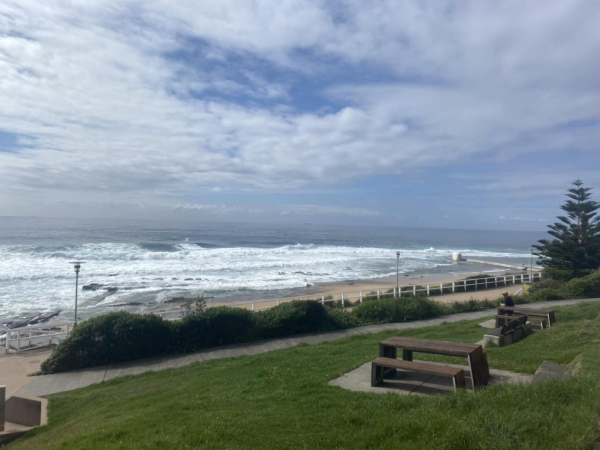 Photo looking over a picnic table and benches on a hill and out past Merewether Beach to the Ocean Baths and  the sea. Green grass on the hill, light sand on the beach, lots of white in the ocean, and a blue sky with white clouds.