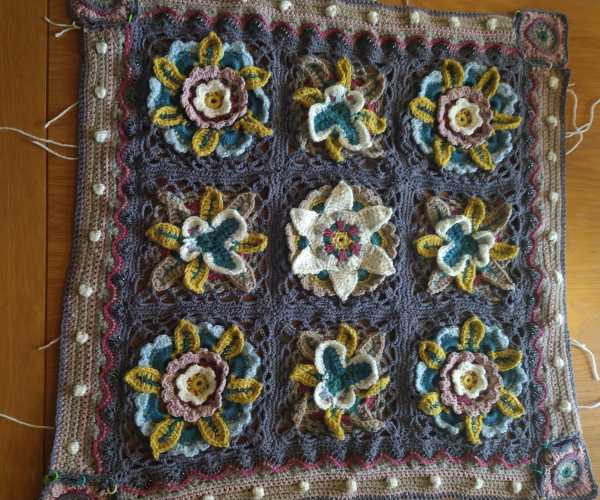 Nine crochet squares with grey background there are four corner flower designs in the centre of each row is a butterfly motif and the central square has a white flower design, the outside border is is in shades of pink with small white bobbles 