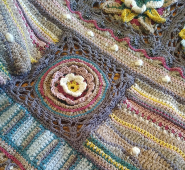 Close up of Jane Crowfoot  crochet Afghan, laid out on a table, it is in greys, blues, pinks, with flower and butterfly motifs, with bobble, wave and bead stitches 