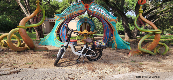 A black and white fat-tire folding ebike in front of an ornate colorful sculpture, part of which looks like an organic archway, with tentacular protrusions around it.