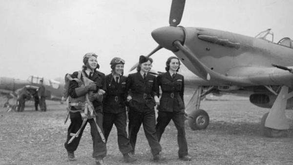 Four smiling Female ATA Air Transport Auxiliary Pilots walk arm in arm across a grassy airfield, in front of a Spitfire fighter plane. Against initial resistance, these young women also flew Hurricanes, and Lancasters. Black and White photo courtesy of the Royal Air Force.