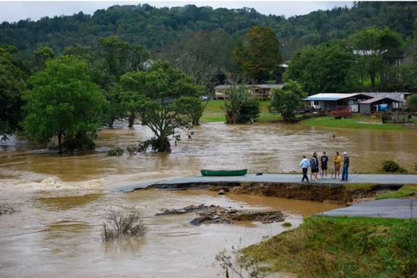 A photograph of flooded areas, with people standing on a washed out road. There are trees in the background, and the water is brown.