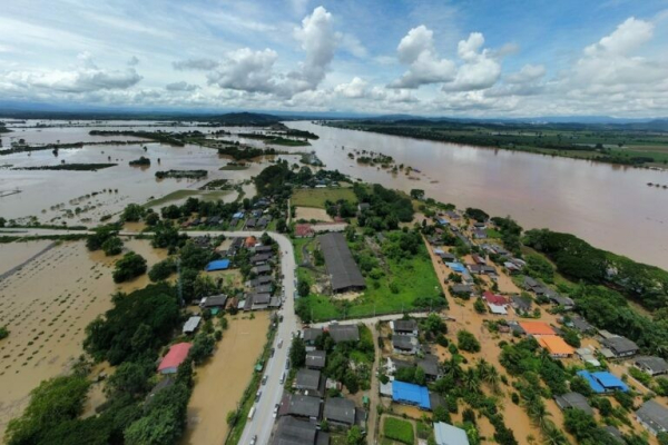 A photograph of a flooded area, with houses and farmland underwater. The sky is mostly blue with a few clouds. The floodwater is brown.