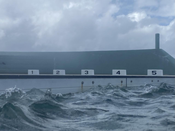 Photo of choppy water hitting the side of the main pool with the neighbouring lap lane blocks and turquoise concrete bleachers in the background. Colours: moody blues with white accents.