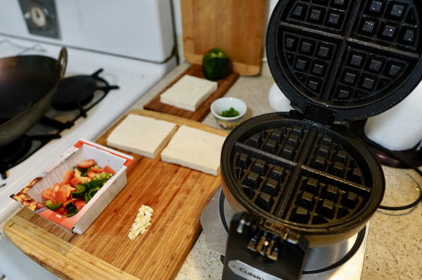 A photo of a kitchen table with a waffle iron, some sliced vegetables, and three pieces of uncooked tofu. The tofu are about 3" by 4" and are about 1/2" tall. 