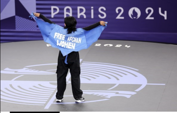 Breakdancer Manizha Talash performs at the Olympics in Paris 2024. She is wearing a cape that looks like wings made from a traditional Afghan blue burka. In white letters are the words Free Afghan Women.