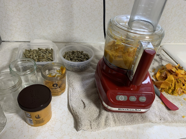 My white kitchen counter showing a red food processer with pumpkin puree, two jars with pumpkin puree, two clear plastic containers full of green pumpkin seeds and a pile of discarded pumpkin skin.