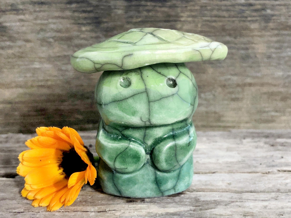 against a background of grey-brown driftwood, and small green nature spirit raku sculpture. it has an open, gently smiling face, two cute little arms, and a pale green leaf on its head. there's an orange calendula flower next to it on the left.