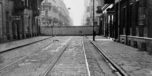 Black and white historical photo of an empty street in the Warsaw Ghetto, featuring tram tracks running down the center, cobblestone pavement, and buildings lining both sides. The wall of the ghetto is visible in the background.