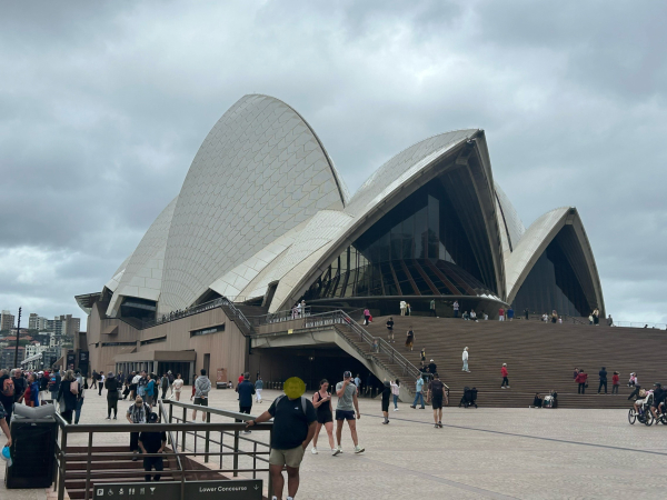 Close view of the landmark Sydney Opera House on a busy Saturday, with many people milling around the surrounding entrance area.  Photo aspect is from the lower left corner and three of the arches and the broad staircase appear prominent - all under a gray sky. A portion of nearby city buildings are seen in the left background. 