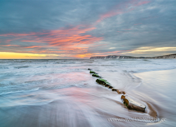 A delicate pink, yellow and blue sunset over the sea and a chalk headland. A narrow rock ledge is visible in the centre of the image, which leads out into the water. A slightly long exposure blurs the sea, but only a little as the texture of the waves remains. A gentle, relaxing view, taken at Compton Bay on the Isle of Wight.