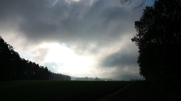 A landscape shot. The foreground of the picture is dark, a path can be faintly made out, with trees at the edge of the forest to the left and right. In the center of the picture there are dense grey clouds that become lighter towards the middle and a milky vague sense can be seen. The mood is gloomy to ominous.  