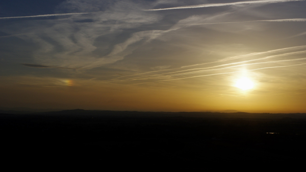 Contrejour vers le sud-ouest, ciel bleu avec une irisation dans les nuages, nuages en zigzag, quelques trainees d'avion qui ressortent avec la lumiere du soleil, soleil pleine face, horizon orange intense contrastant avec quelques series de collines a contre-jour. On apercoit le mont valier qui sort des nuages