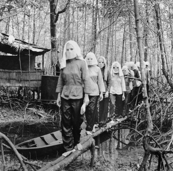 A line of women dressed in dark clothing and white head coverings walks along a narrow wooden bridge over shallow water in a mangrove environment. Nearby, a traditional structure can be seen partially obscured in the dense foliage. The scene is captured in black and white, enhancing the somber atmosphere.